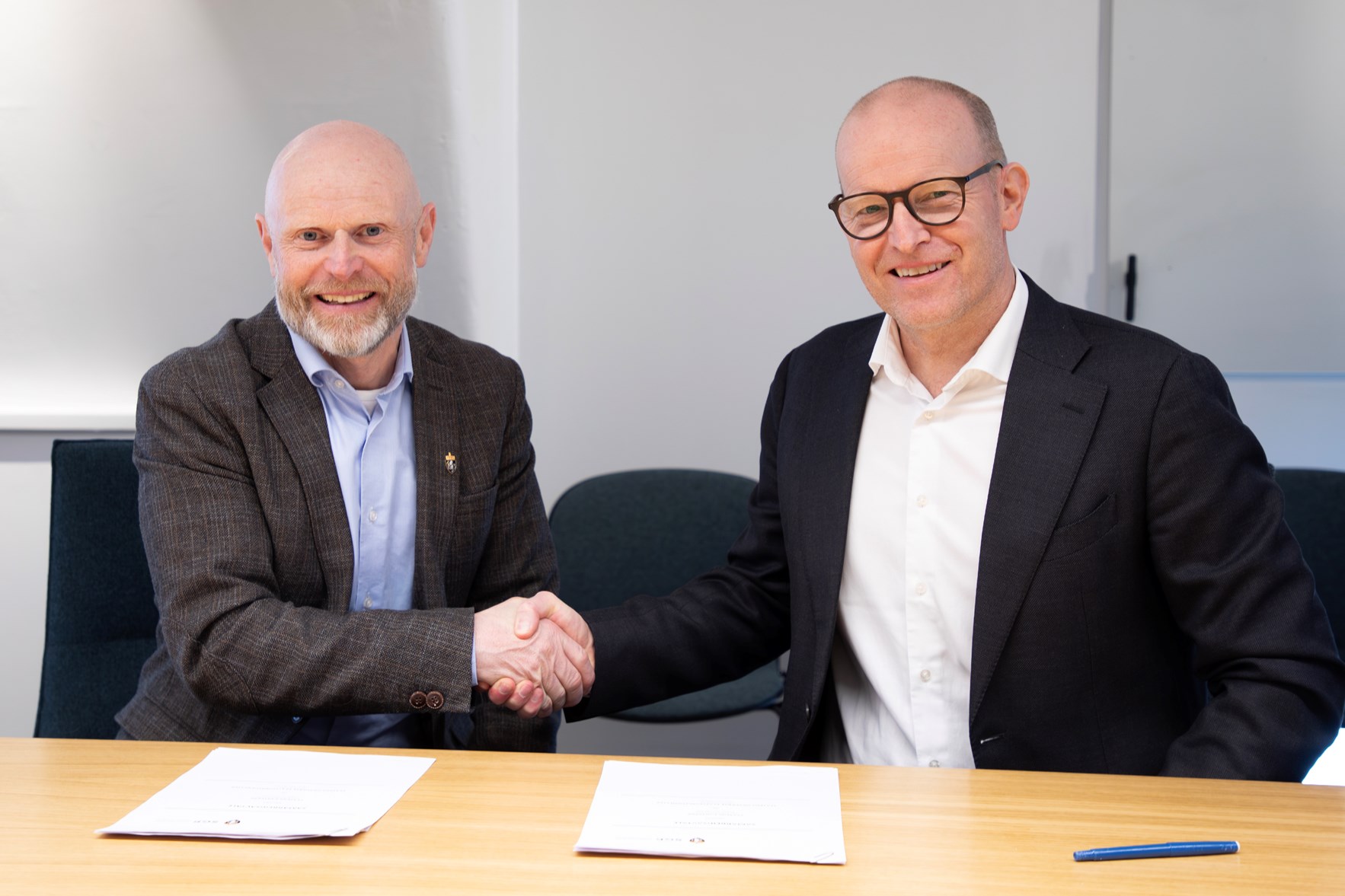 To menn i dress h&aring;ndhilser over et bord med dokumenter.