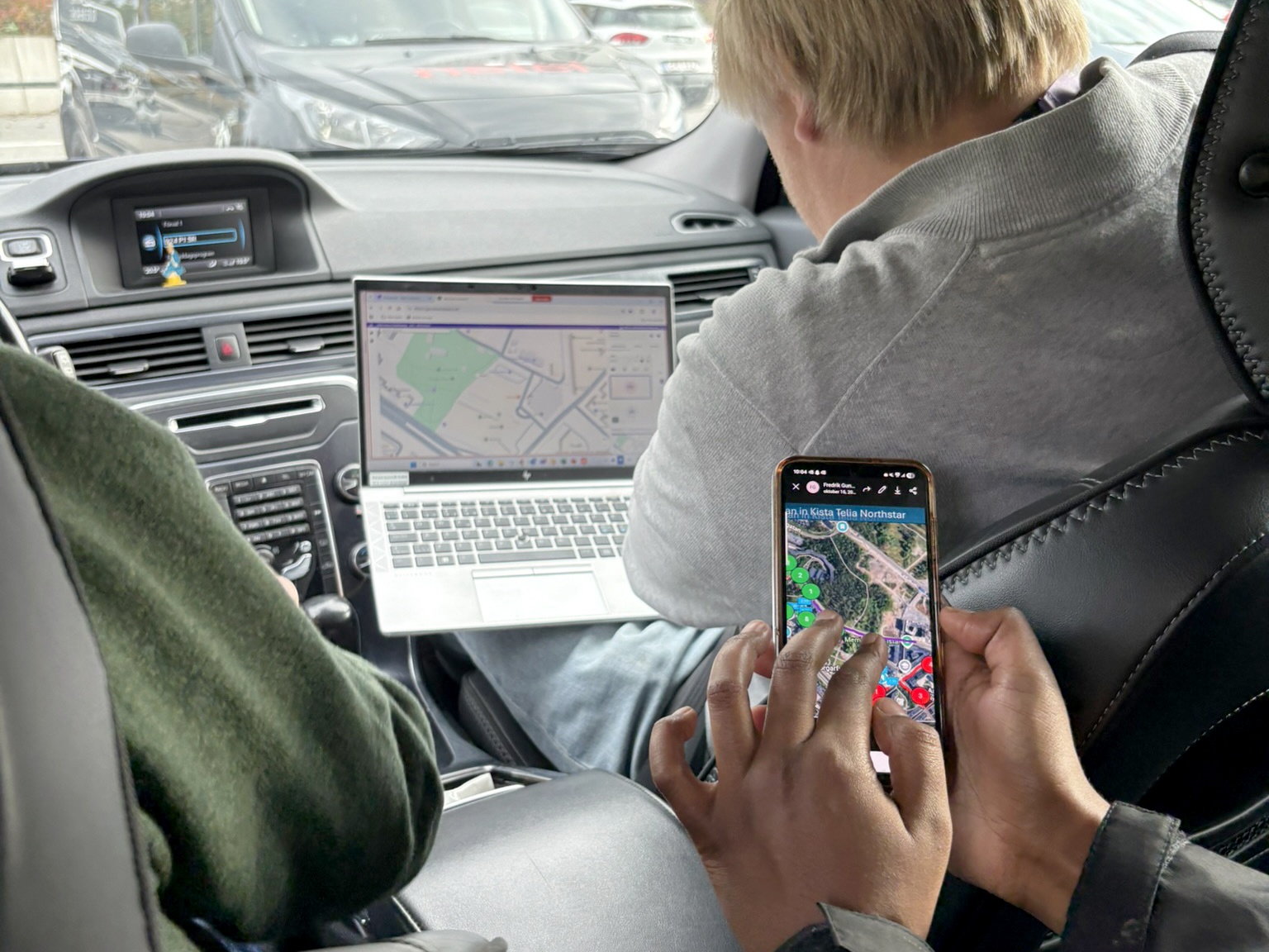 En person bruker en bærbar PC med kart på skjermen i baksetet av en bil, mens en annen holder en mobil med et kart på skjermen. Sjåføren ser på veien.