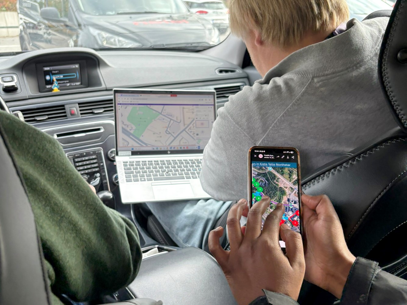 En person bruker en bærbar PC med kart på skjermen i baksetet av en bil, mens en annen holder en mobil med et kart på skjermen. Sjåføren ser på veien.
