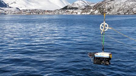 En undervannsfarkost henger over vannflaten og skal senkes ned til havbunnen. Utsikt til snøkledte fjell og blå himmel over bebyggelse på Skjervøy.