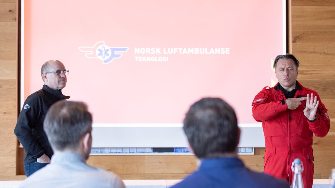To personer presenterer foran en skjerm med logoen til Norsk Luftambulanse. En av dem er kledd i rød uniform. Publikum sitter med ryggen til kameraet.