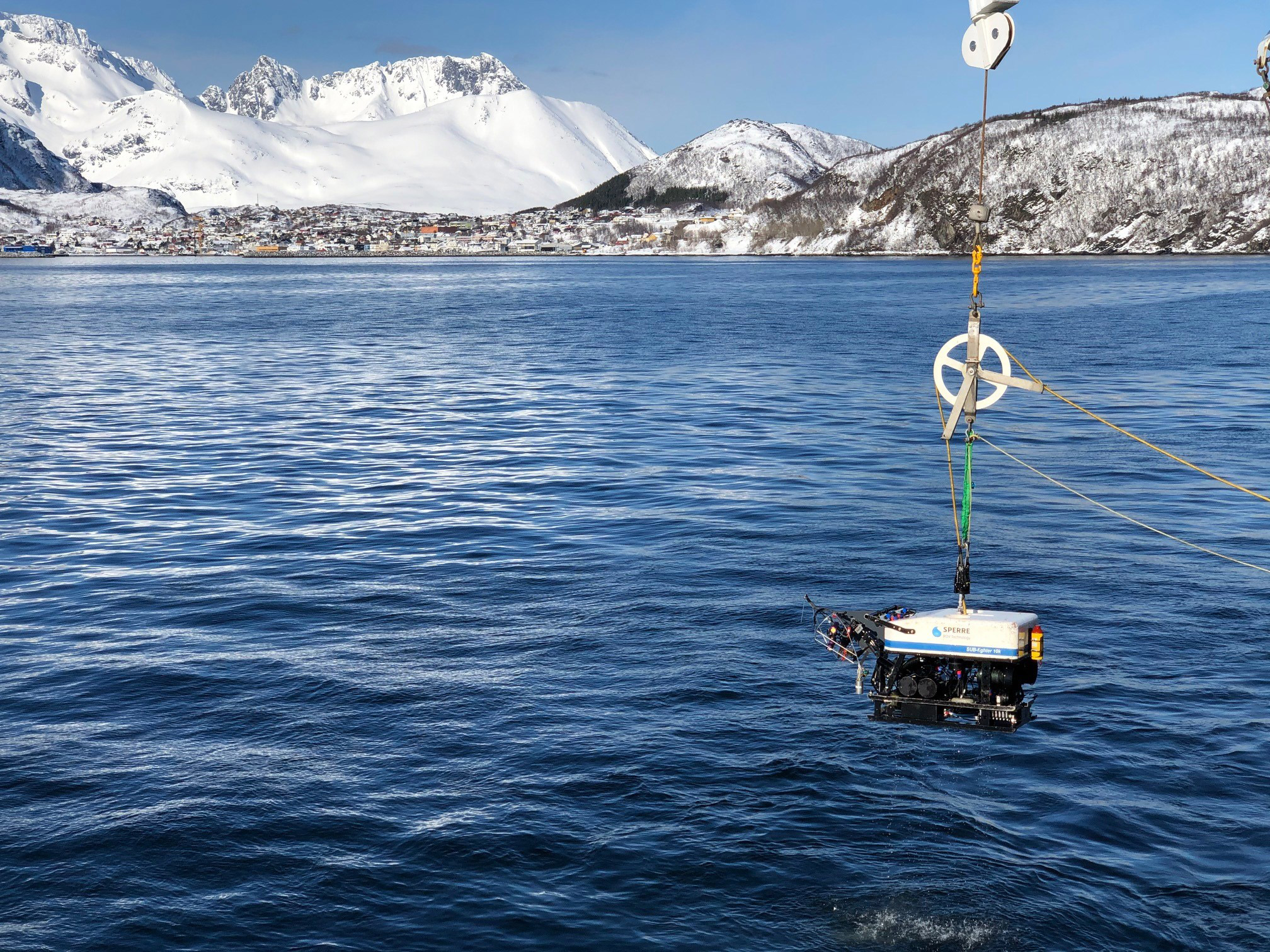 En undervannsfarkost henger over vannflaten og skal senkes ned til havbunnen. Utsikt til sn&oslash;kledte fjell og bl&aring; himmel over bebyggelse p&aring; Skjerv&oslash;y.