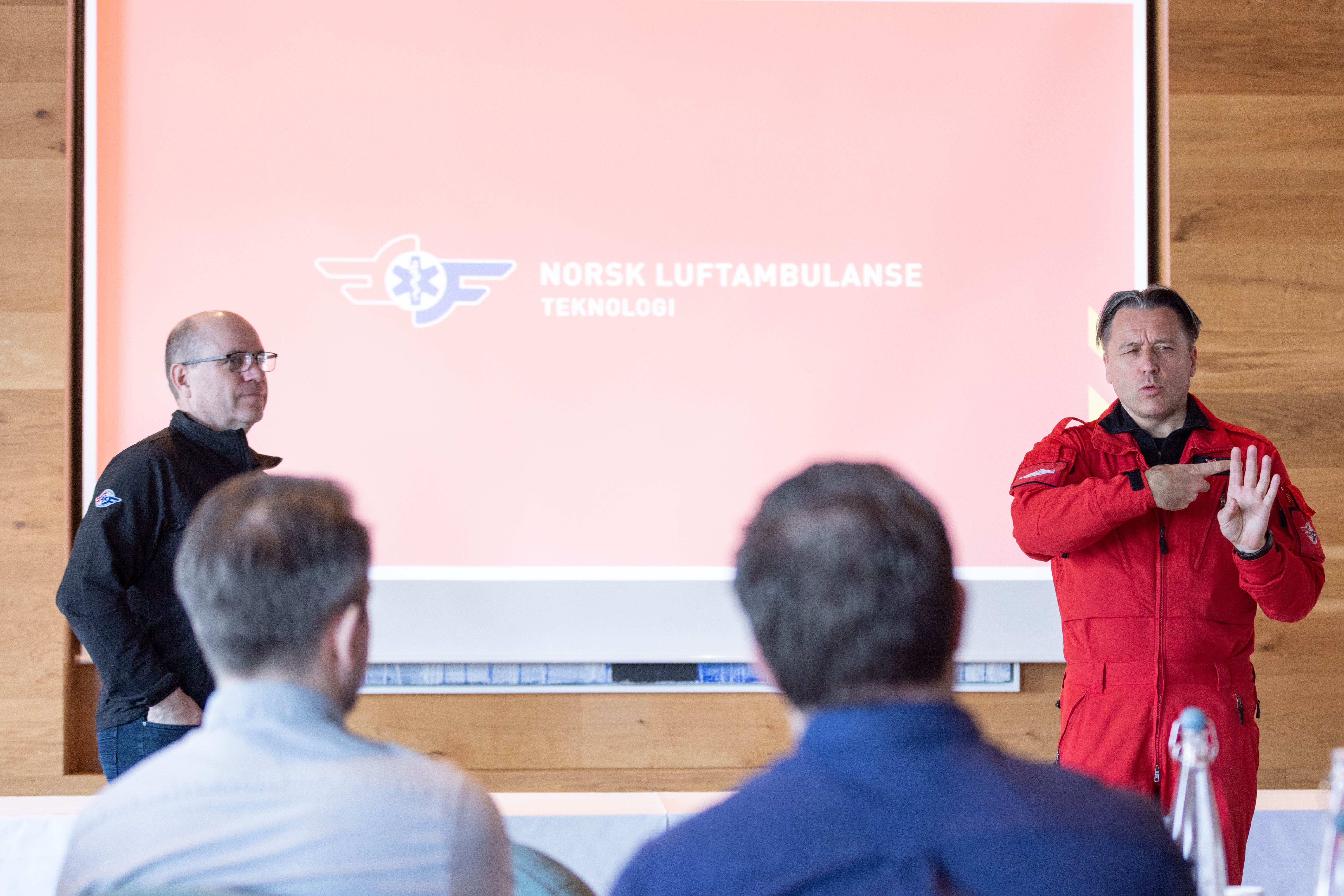 To personer presenterer foran en skjerm med logoen til Norsk Luftambulanse. En av dem er kledd i r&oslash;d uniform. Publikum sitter med ryggen til kameraet.