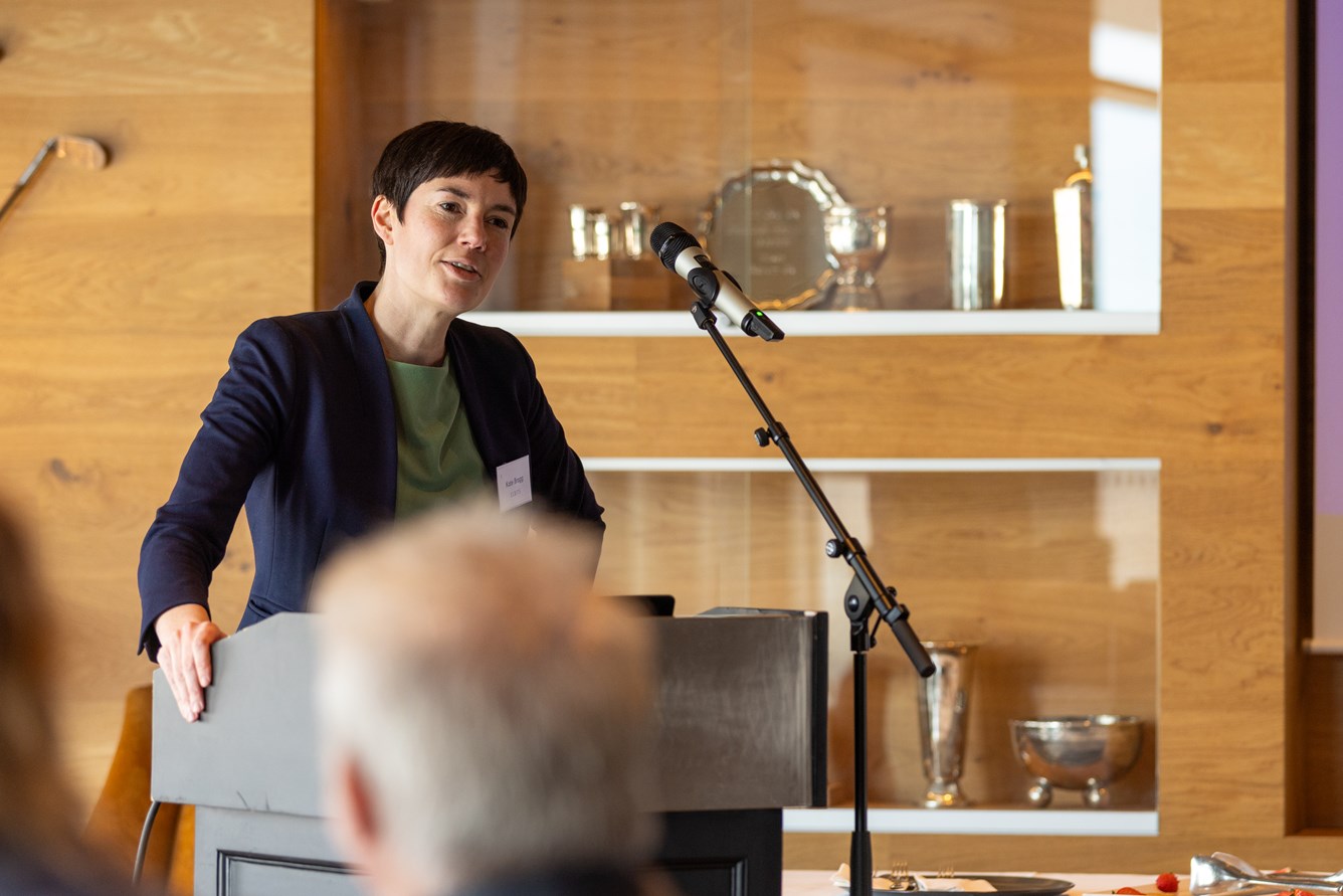 En person holder en tale ved en talerstol med mikrofon. Bakgrunnen har trehyller med diverse gjenstander.