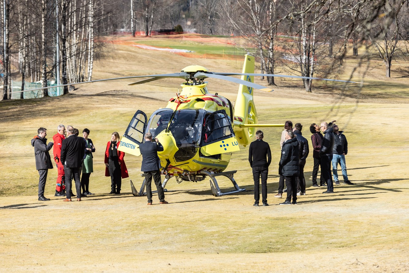 En gruppe mennesker st&aring;r rundt et gult helikopter fra Norsk Luftambulanse som st&aring;r p&aring; en gressplen.