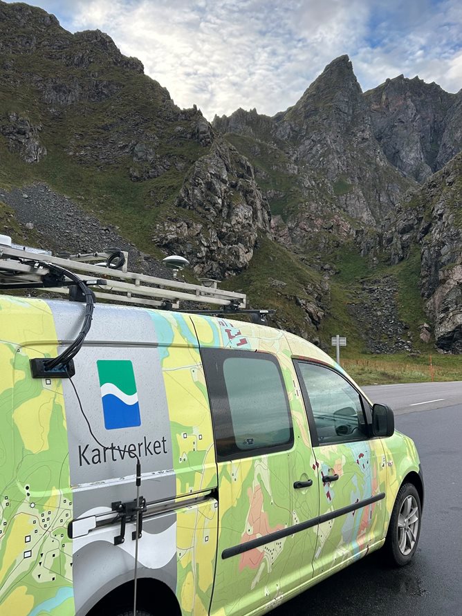 Varebil med Kartverket-logo og teknisk utstyr på taket foran bratt fjell.