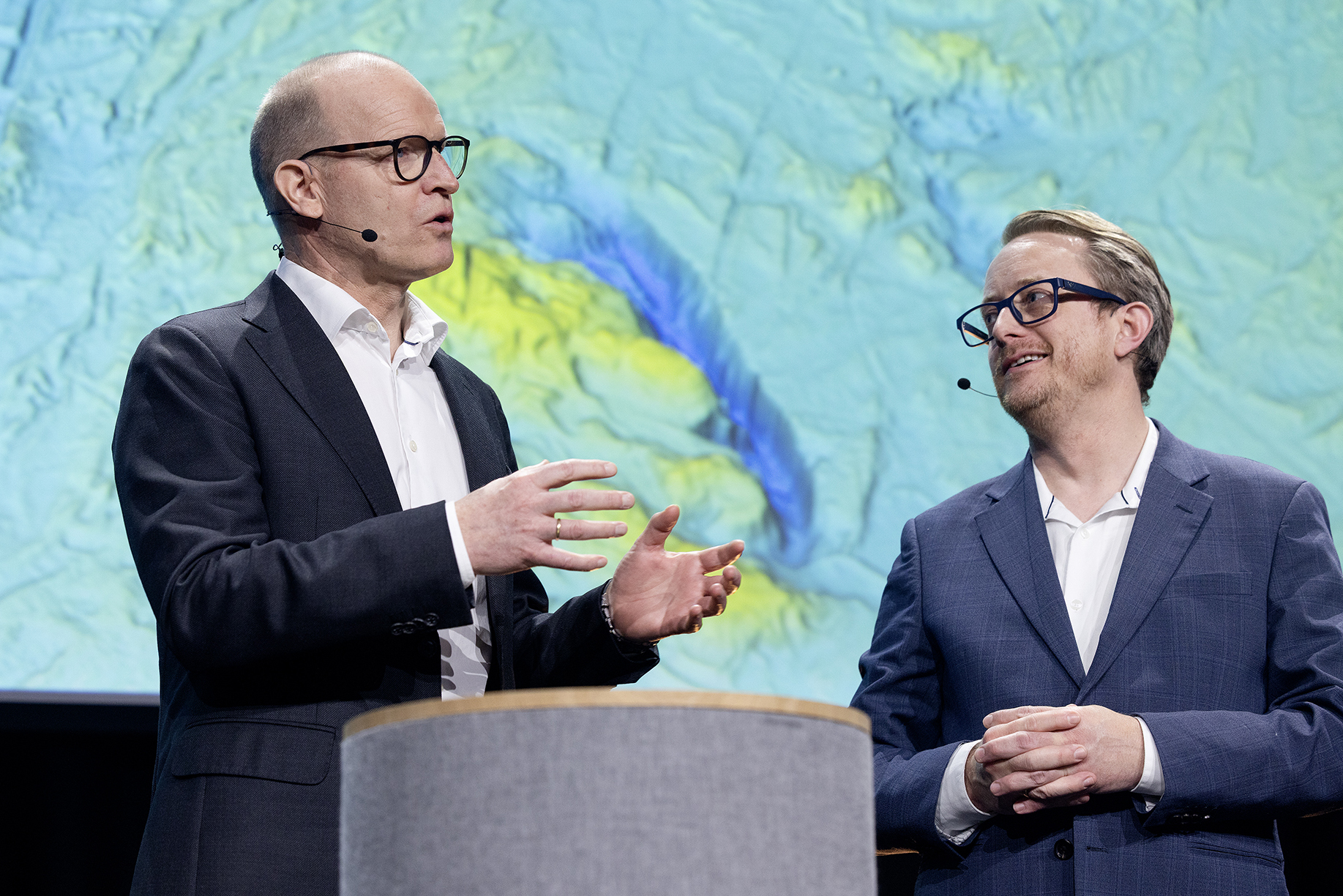 To dresskledte menn på en scene. De virker engasjerte og gestikulerer ivrig.