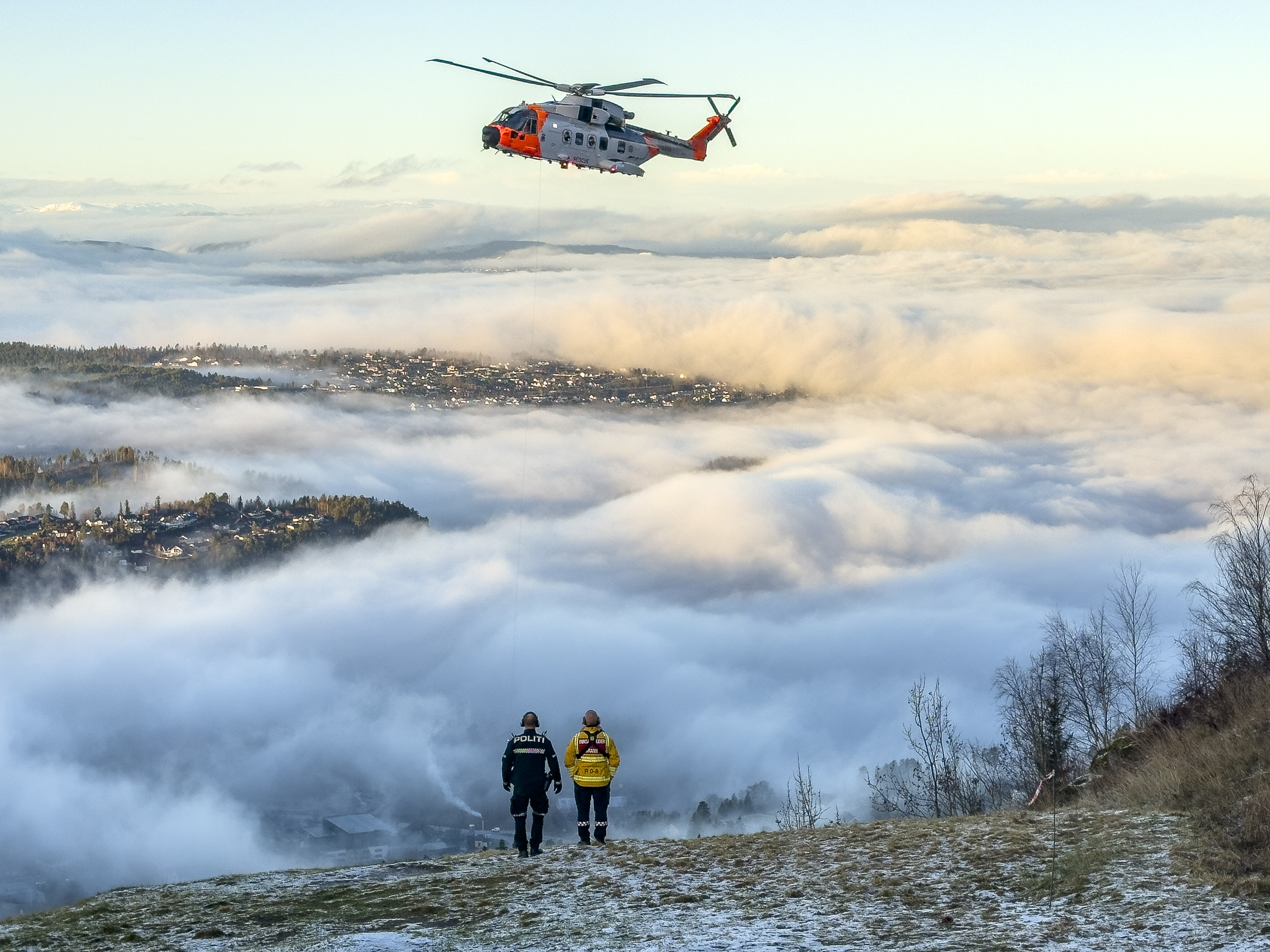 Et redningshelikopter som svever i en bratt skråning med hus i tåke under. En politimann og en brannmann står med ryggen til.