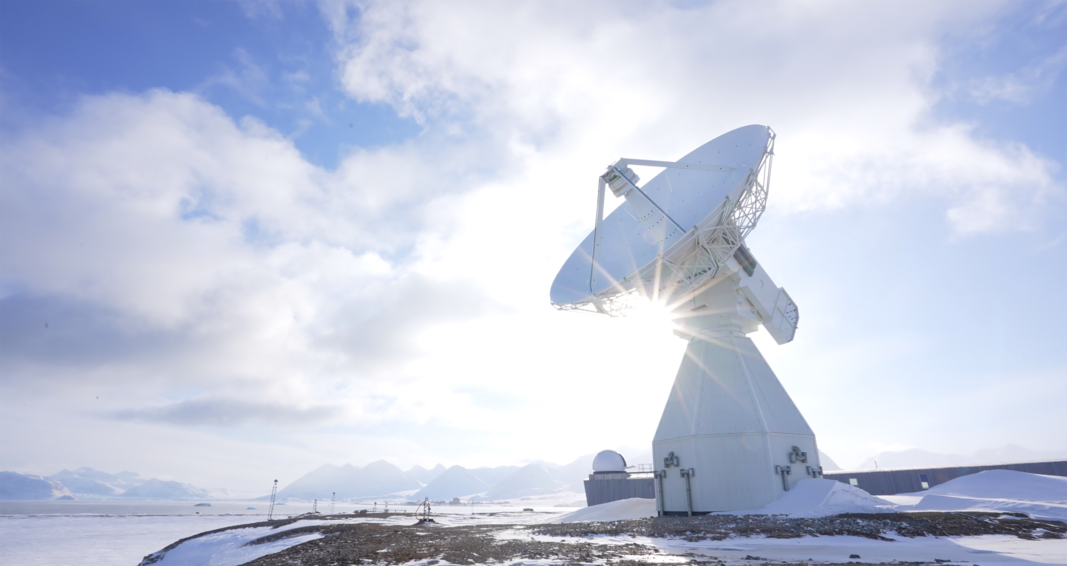 En av Kartverkets satellittantenner ved jordobservatoriet i Ny-Ålesund. Et høyt, hvitt tårn med en parabol-disk fotografert i motlys mot snødekte fjell og en fjord.