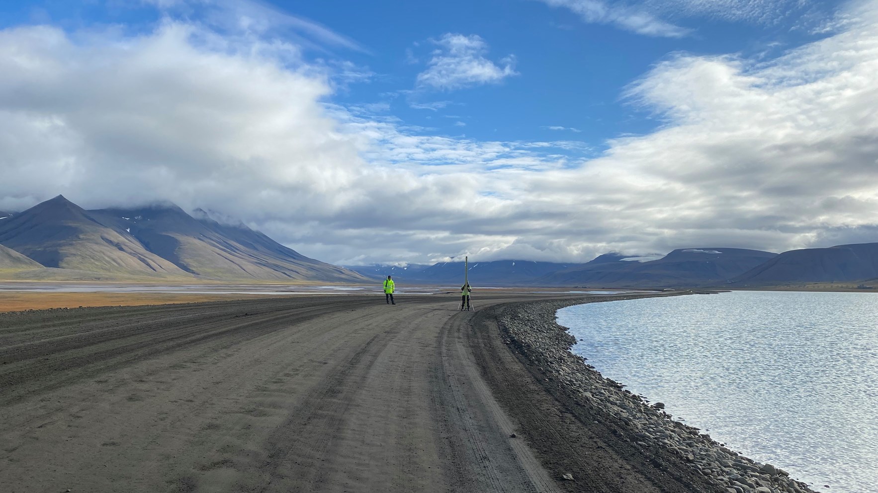 To personer i gule jakker utfører feltarbeid langs en grusvei ved siden av hav, med fjellandskap i bakgrunnen under en skyet himmel.