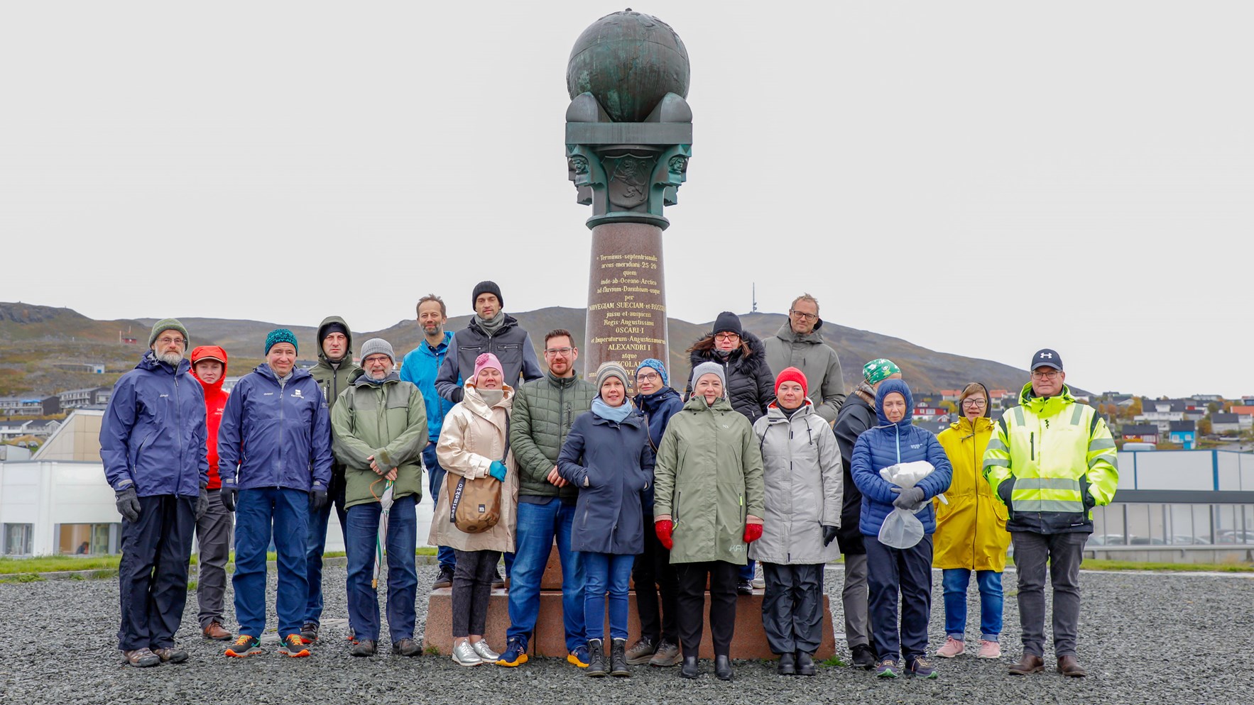 En gruppe mennesker står samlet foran meridianstøtten i Hammerfest.
