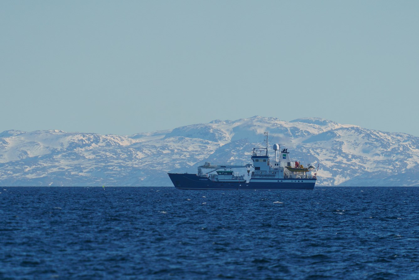 Bilde av en stor b&aring;t med fjord og fjell i bakgrunnen.
