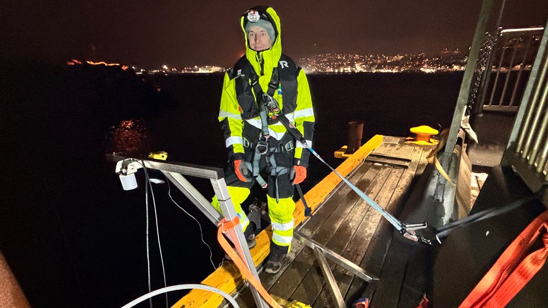 Mann i arbeidstøy, stroppet fast, står ved en vannstandsmåler på ei kai.