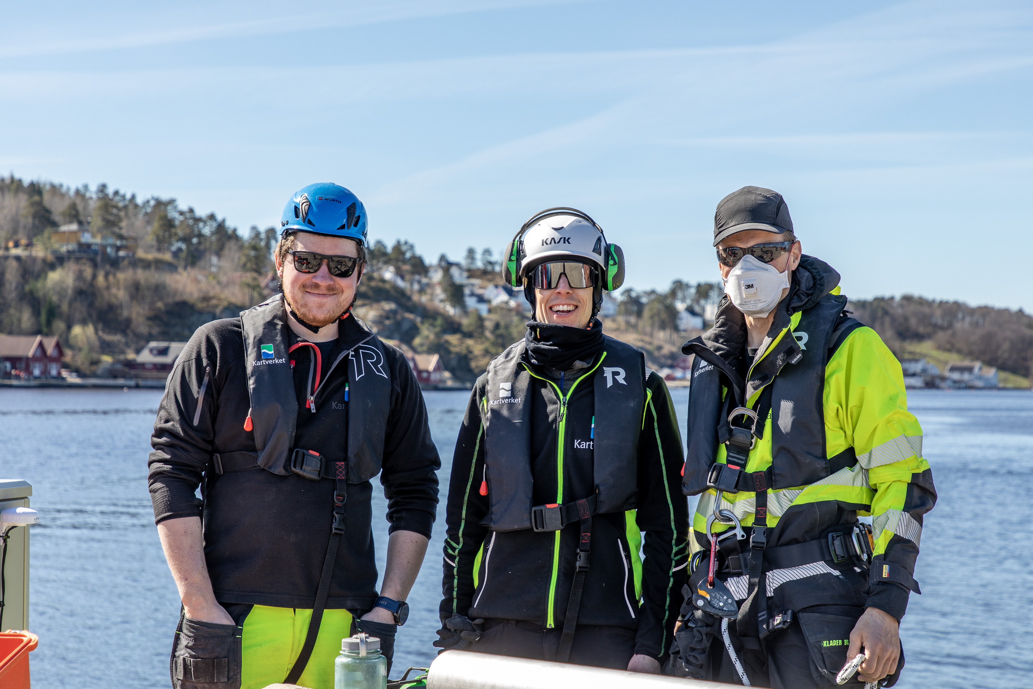 Tre mannlige arbeidere med hjelmer, solbriller og signalbekledning smiler mot kamera. I bakgrunnen ser vi sjø og sørlandsk kystnatur.