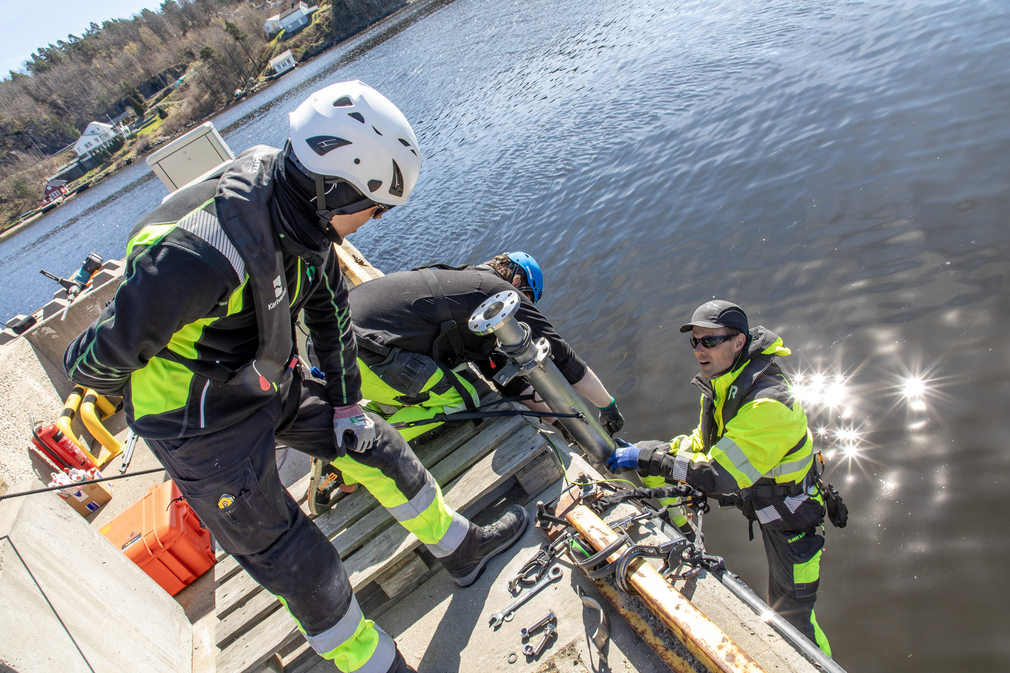 To personer st&aring;r og jobber p&aring; en kaikant mens den tredje st&aring;r i en stige som henger ned fra bryggekanten, mens de monterer teknisk utstyr.